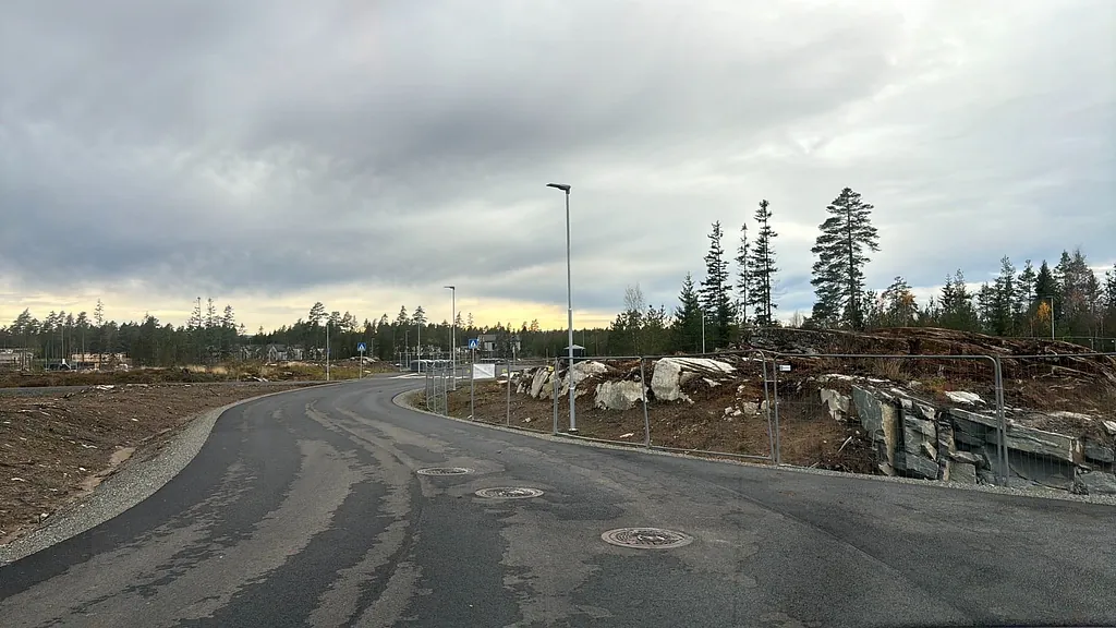 Nyasfaltert vei med gatelys og midlertidige gjerder slynger seg gjennom byggefelt omgitt av skog og stein.