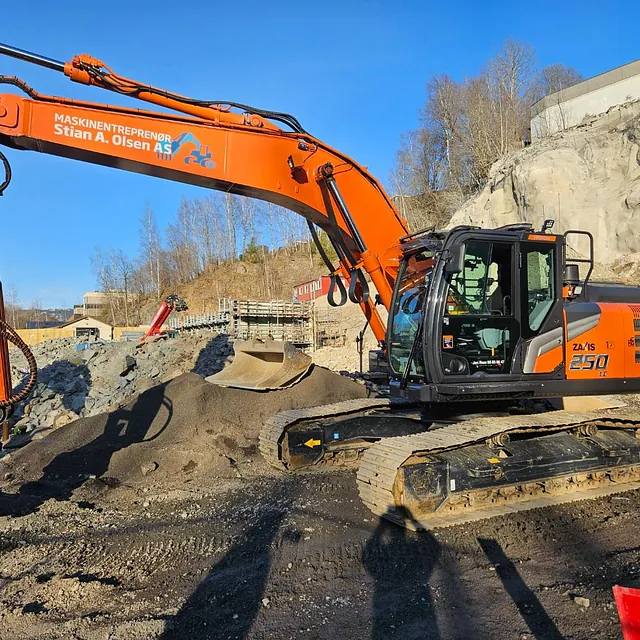 Gravemaskin jobber med masser på en steinete tomt med fjellvegg og blå himmel i bakgrunnen.