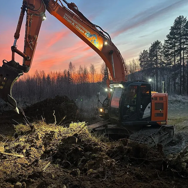 Gravemaskin løfter jord i frostdekket terreng ved soloppgang med fargerik himmel og trær.