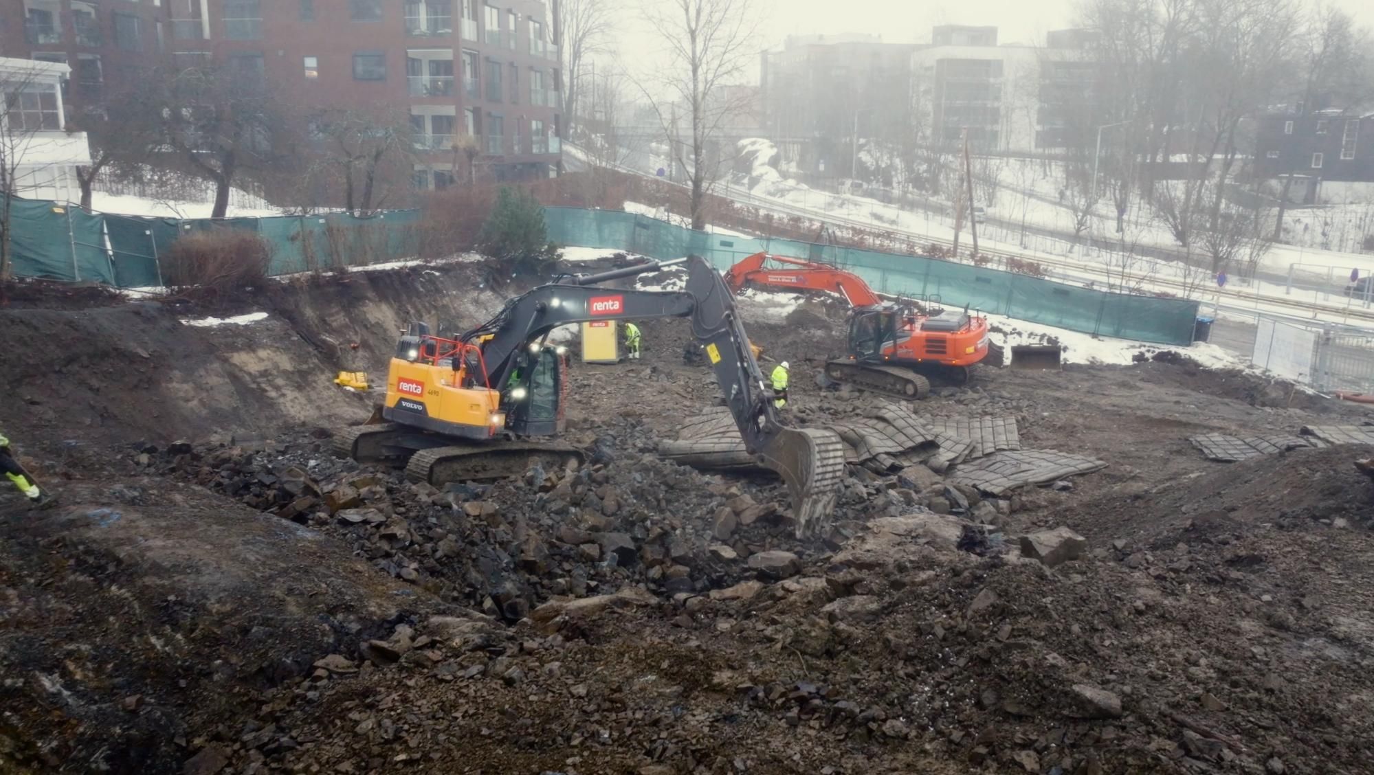 Gravemaskiner og arbeidere i stor byggegrop med sprengstein og masser