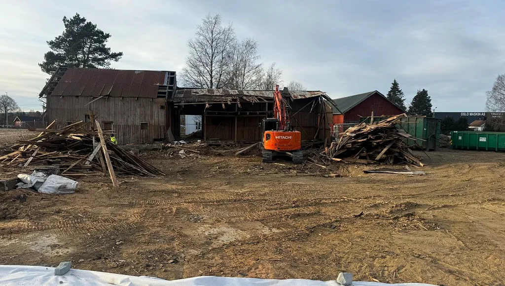 Rivningsarbeid med gravemaskin foran en gammel låve, omgitt av rivningsmaterialer og avfallscontainere.