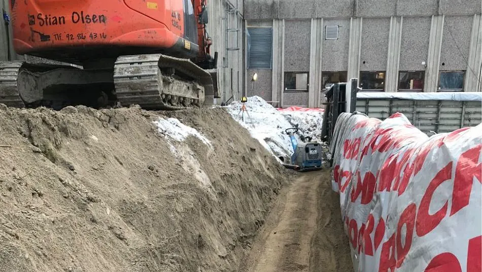 Gravemaskin står ved grøft fylt med sand inntil byggvegg.
