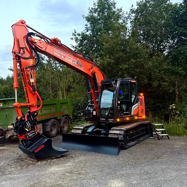Oransje gravemaskin med skuff står parkert på grusplass ved trær og tilhenger i bakgrunnen.