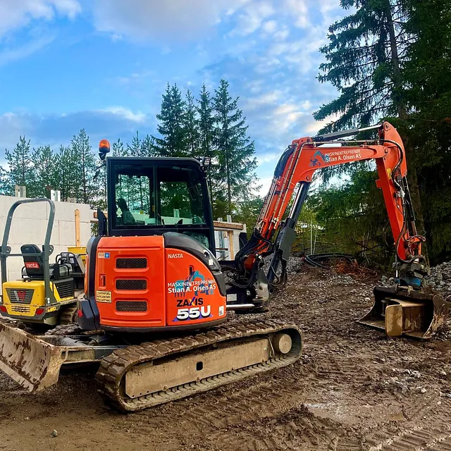 Oransje minigraver med Stian A. Olsen AS-logo står på en gjørmete byggeplass foran furutrær.