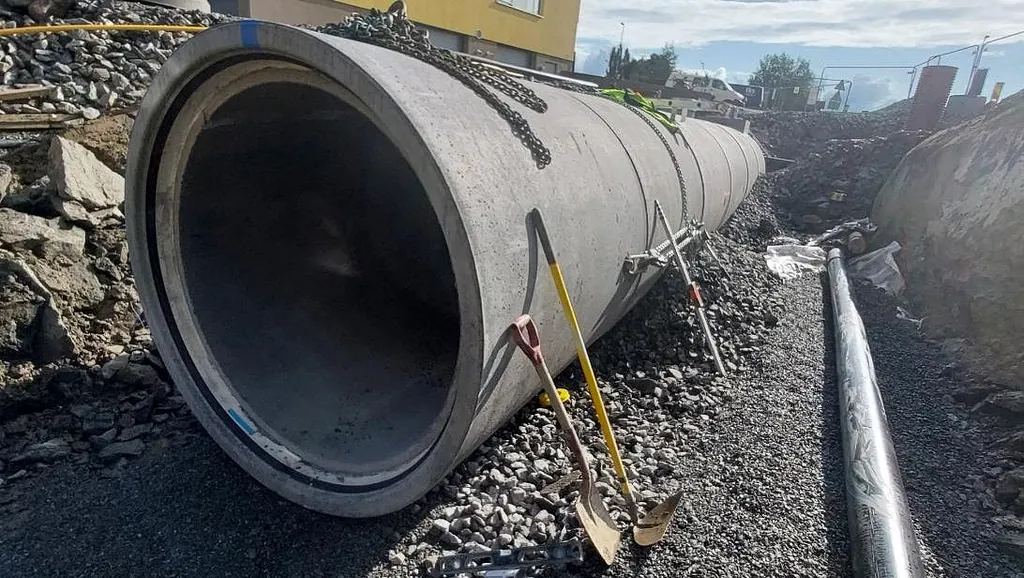 Betongrør og verktøy ligger klart på byggeplass med gul bygning og blå himmel i bakgrunnen.