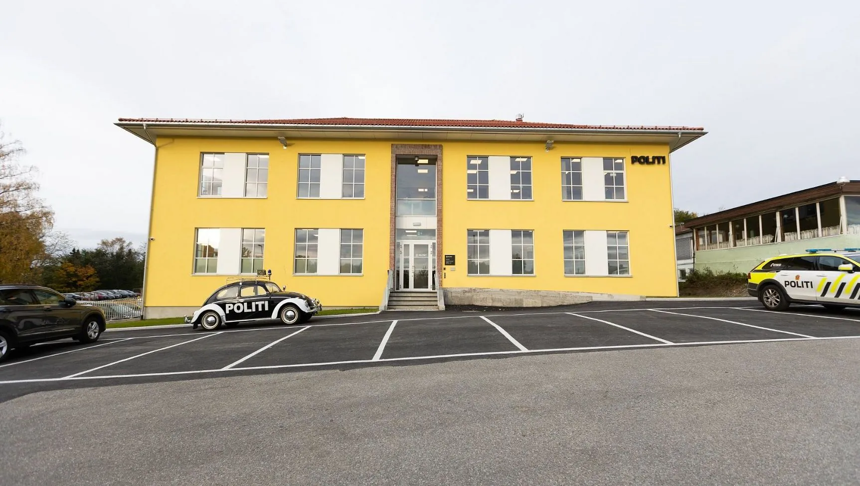 Gul politistasjon med parkeringsplass foran, biler og en svart-hvit politibil utenfor.