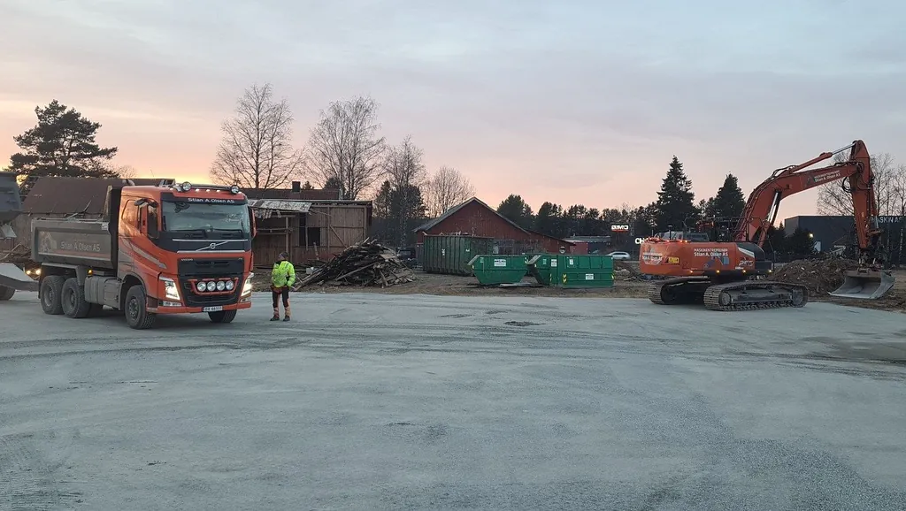 Oransje lastebil og gravemaskin i arbeid ved riving, med containere og trehus i bakgrunnen.
