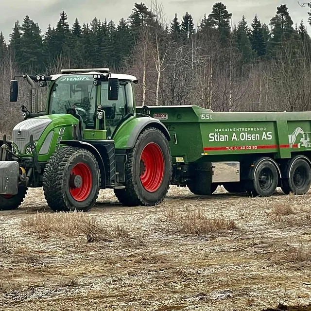 Grønn Fendt-traktor med tilhenger fra Stian A. Olsen AS står på en åker med skog i bakgrunnen.