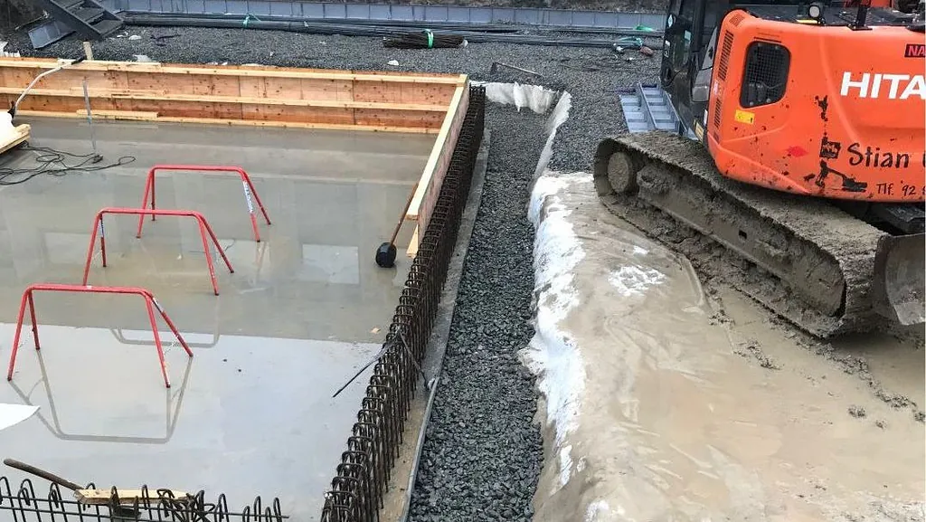 Gravemaskin ved støpt betongfundament med armering og vann på byggeplass.