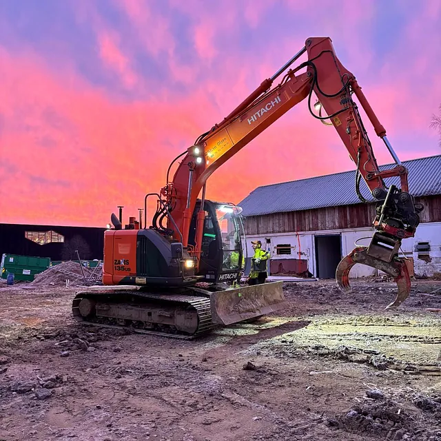Gravemaskin med tømmerklo i arbeid foran en låve ved soloppgang med rosa og lilla himmel.