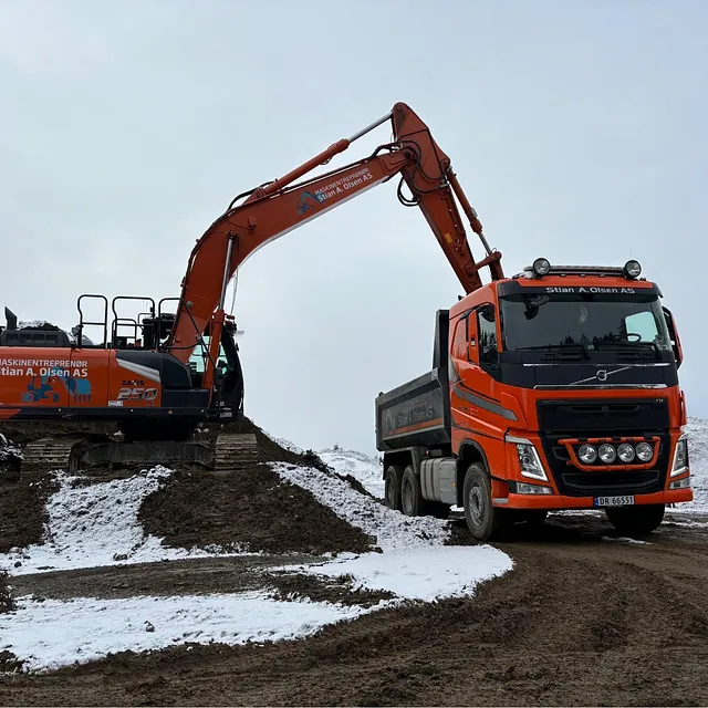 Gravemaskin laster jord i lastebil fra Stian A. Olsen AS på snødekt byggeplass i gråvær.