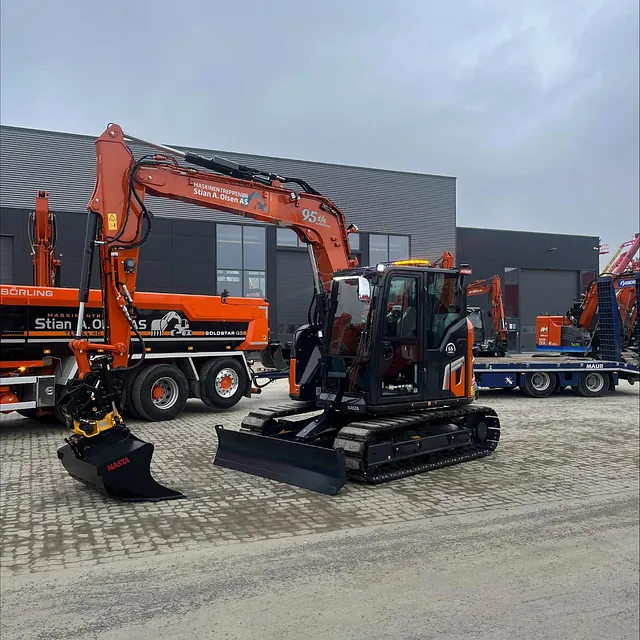 Ny gravemaskin parkert foran verksted med lastebil og maskiner i bakgrunnen