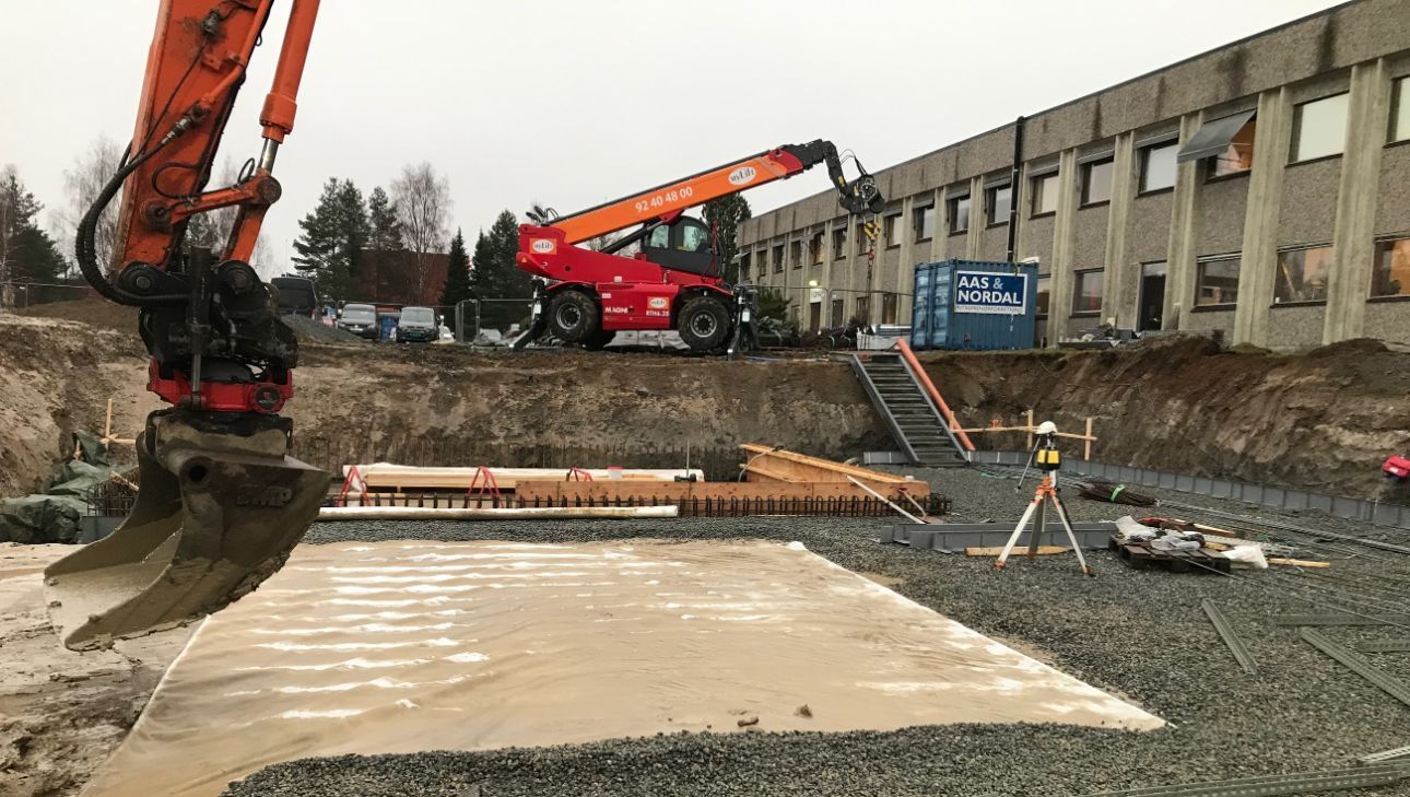 Gruslagt byggegrop med armeringsjern, gravemaskin og teleskoplaster bak.