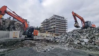 To gravemaskiner flytter stein på byggeplass foran blokk under oppføring.