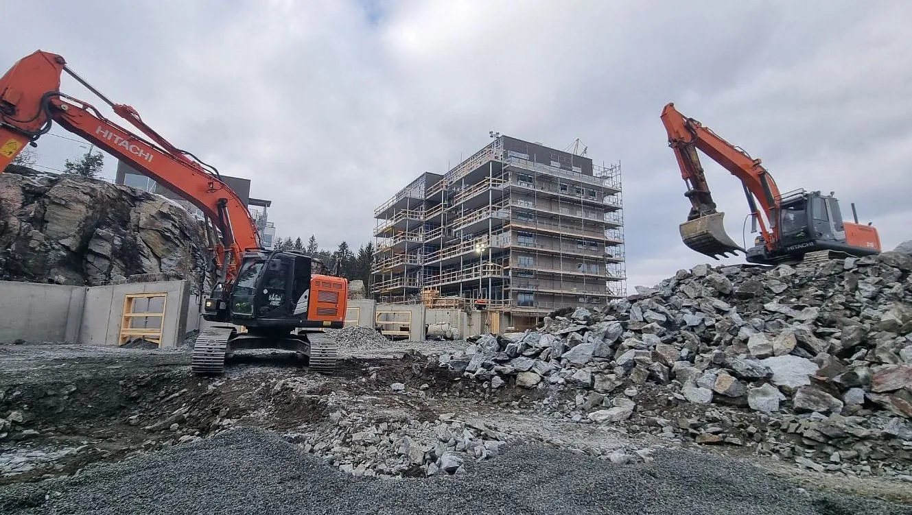 To gravemaskiner flytter stein på byggeplass foran blokk under oppføring.