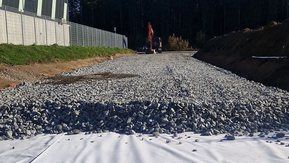 Underlag med hvit duk og grov stein legges ut ved industriområde, gravemaskin i bakgrunnen nær skogkant.