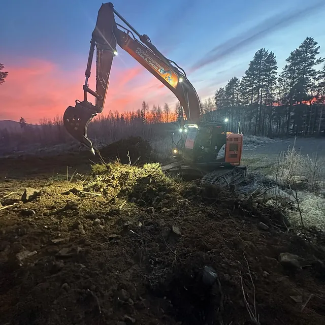 Gravemaskin lyser opp jordhauger i mørketid med oransje og blå himmel over frostdekket landskap.