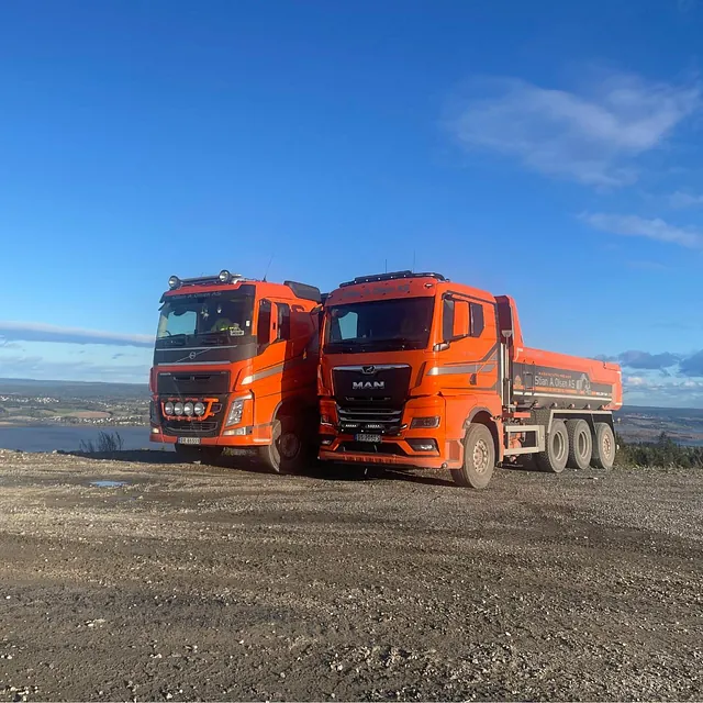 To dumpere parkert på grusplass med utsikt over landskap og vann
